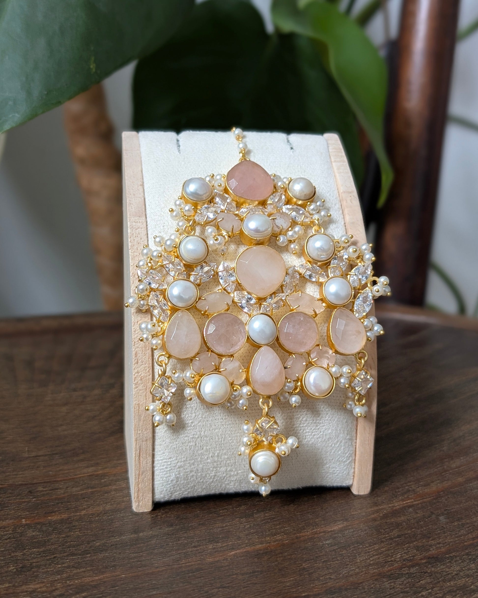 A wide gold-toned cuff bracelet decorated with blush pink rose quartz stones, white faux pearls, and clear crystals, displayed on a cream-colored jewelry stand on a wooden table with a plant in the background.