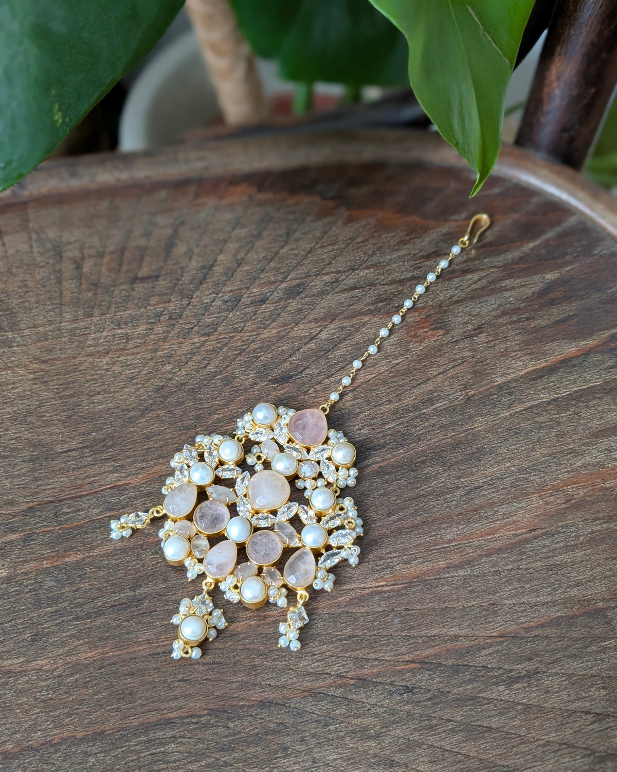 A gold-toned traditional Indian maang tikka with pinkish rose quartz stones and white faux pearls, displayed on a dark wooden surface with green leaves in the background.