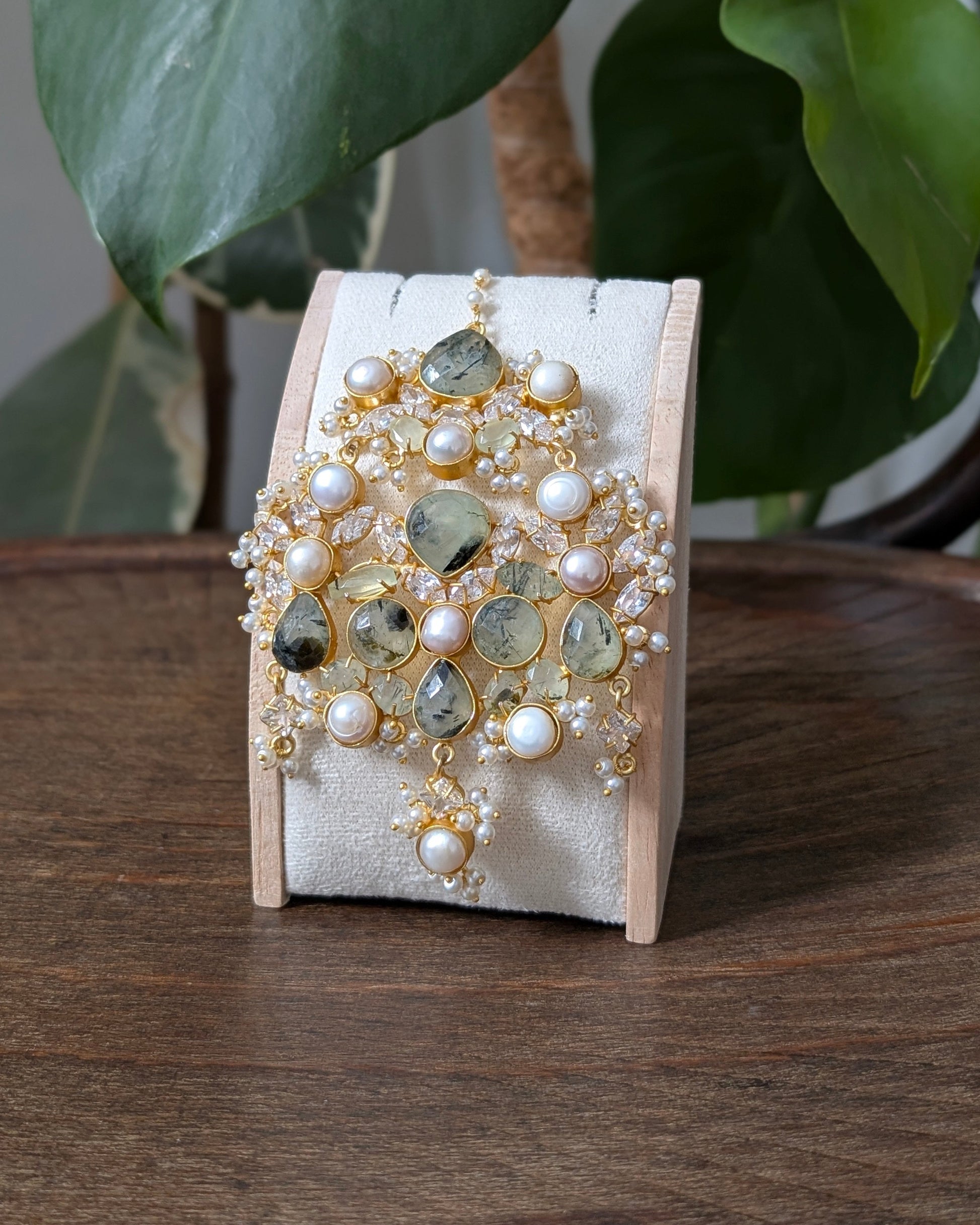 A gold and green gemstone brooch with white pearls, displayed on a cream velvet jewelry stand on a dark wooden table, with green houseplants in the background.