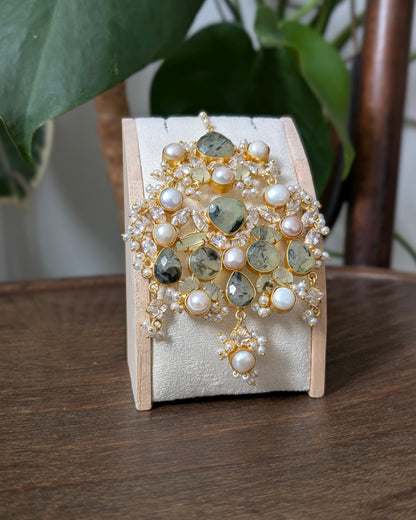 A gold-toned statement brooch featuring natural greenish-yellow teardrop stones, white faux pearls, and small clear crystal accents, displayed on a cream velvet jewelry stand on a wooden table with a plant in the background.