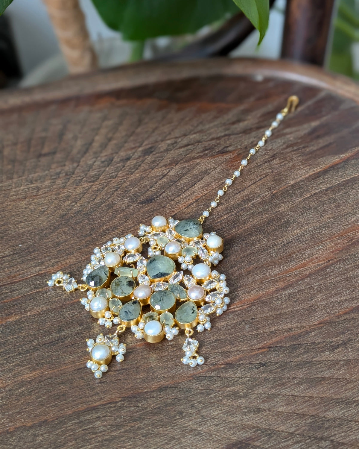 A gold-plated Indian style maang tikka with green stones and white pearls, displayed on a dark wooden surface.