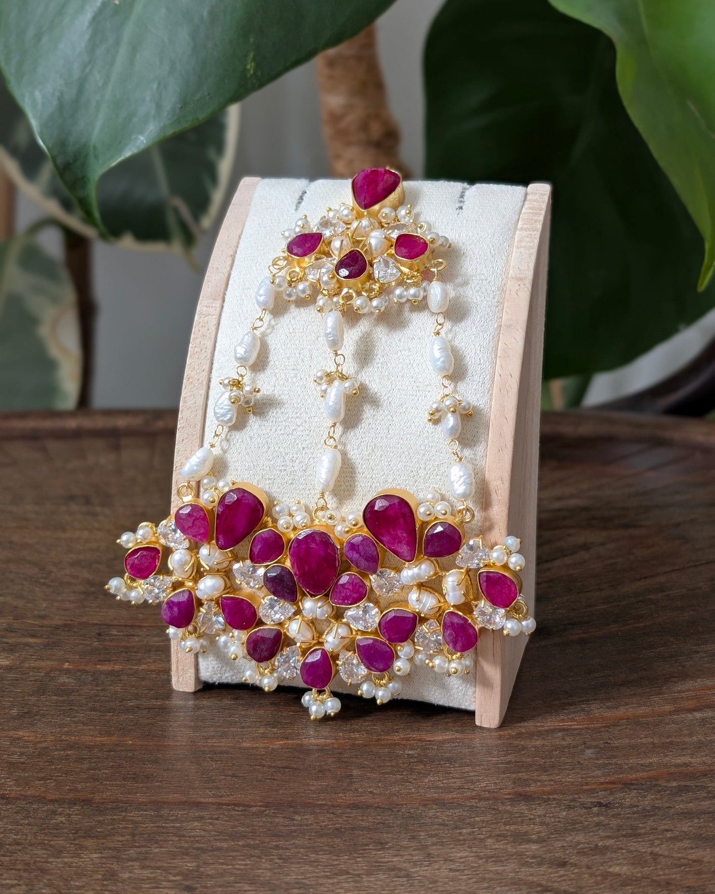 Gold and white pearl traditional Indian jewelry set, including a maang tikka and matching earrings, displayed on a wooden stand on a wooden table with green leaves in the background.