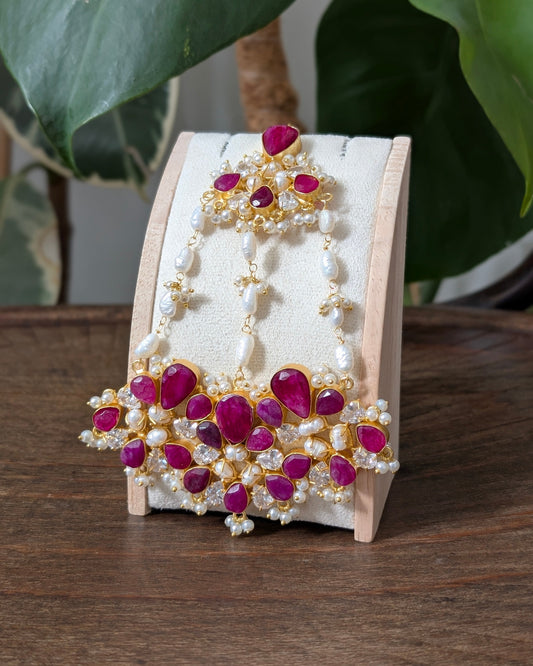 Gold and white pearl traditional Indian jewelry set, including a maang tikka and matching earrings, displayed on a wooden stand on a wooden table with green leaves in the background.