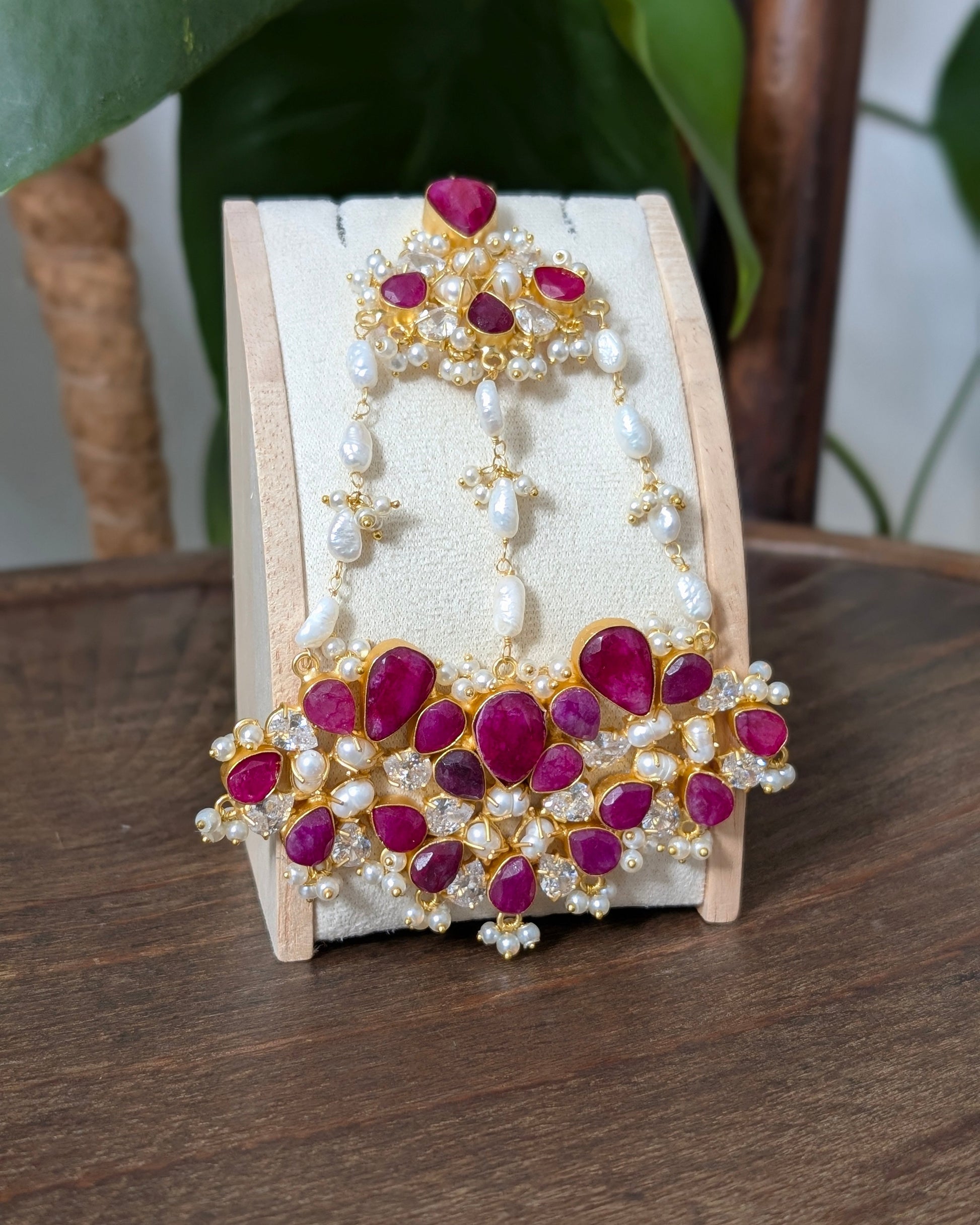 Gold-plated Indian bridal jewelry set featuring a matching headpiece and collar, embellished with deep pinkish-red stones and white pearls, displayed on a cream-colored stand.