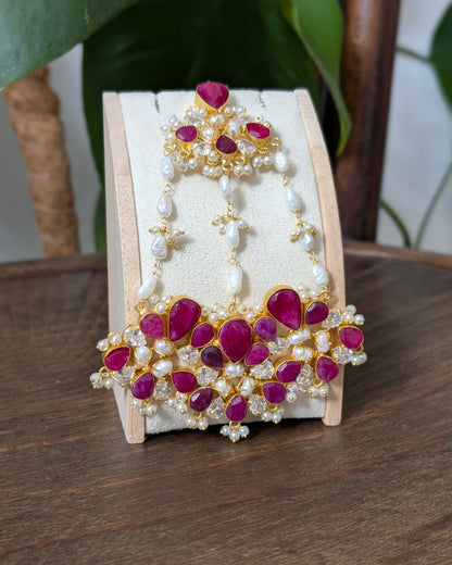 Gold-plated Indian bridal jewelry set featuring a matching headpiece and collar, embellished with deep pinkish-red stones and white pearls, displayed on a cream-colored stand.