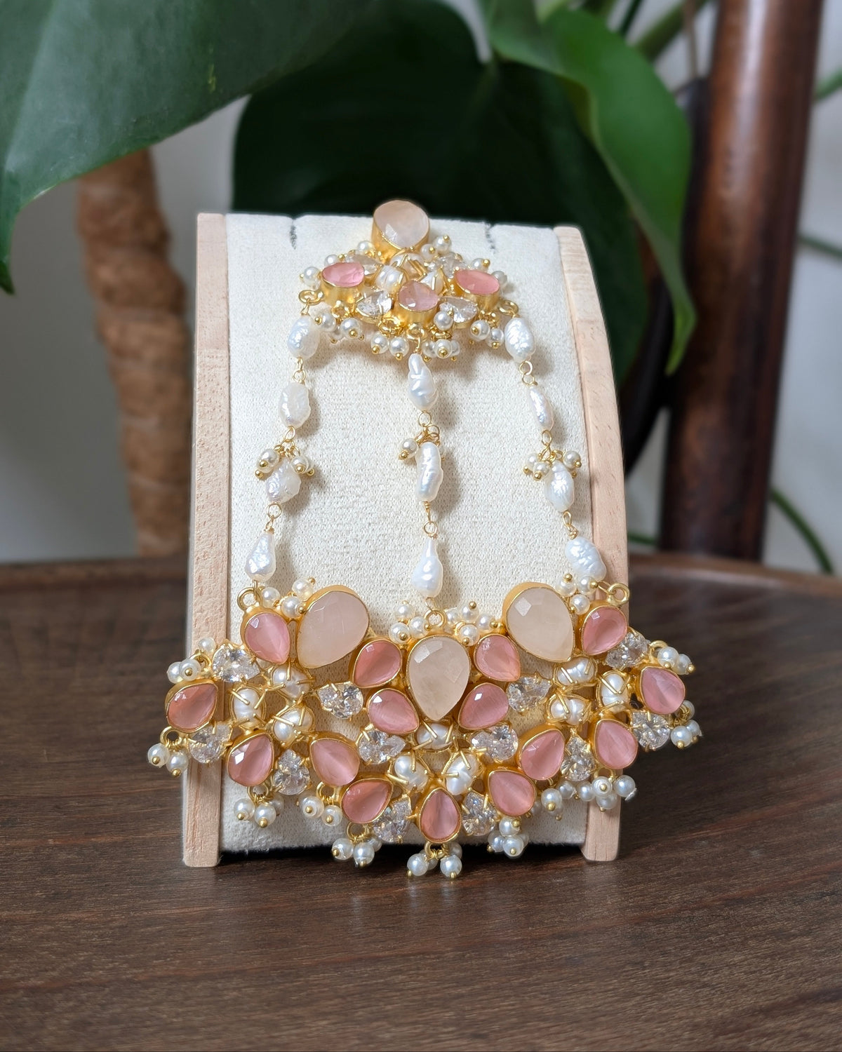 An ornate gold-toned traditional Indian jewelry set featuring pink stone and pearl elements, displayed on a wooden stand on a wooden table with a green plant in the background.