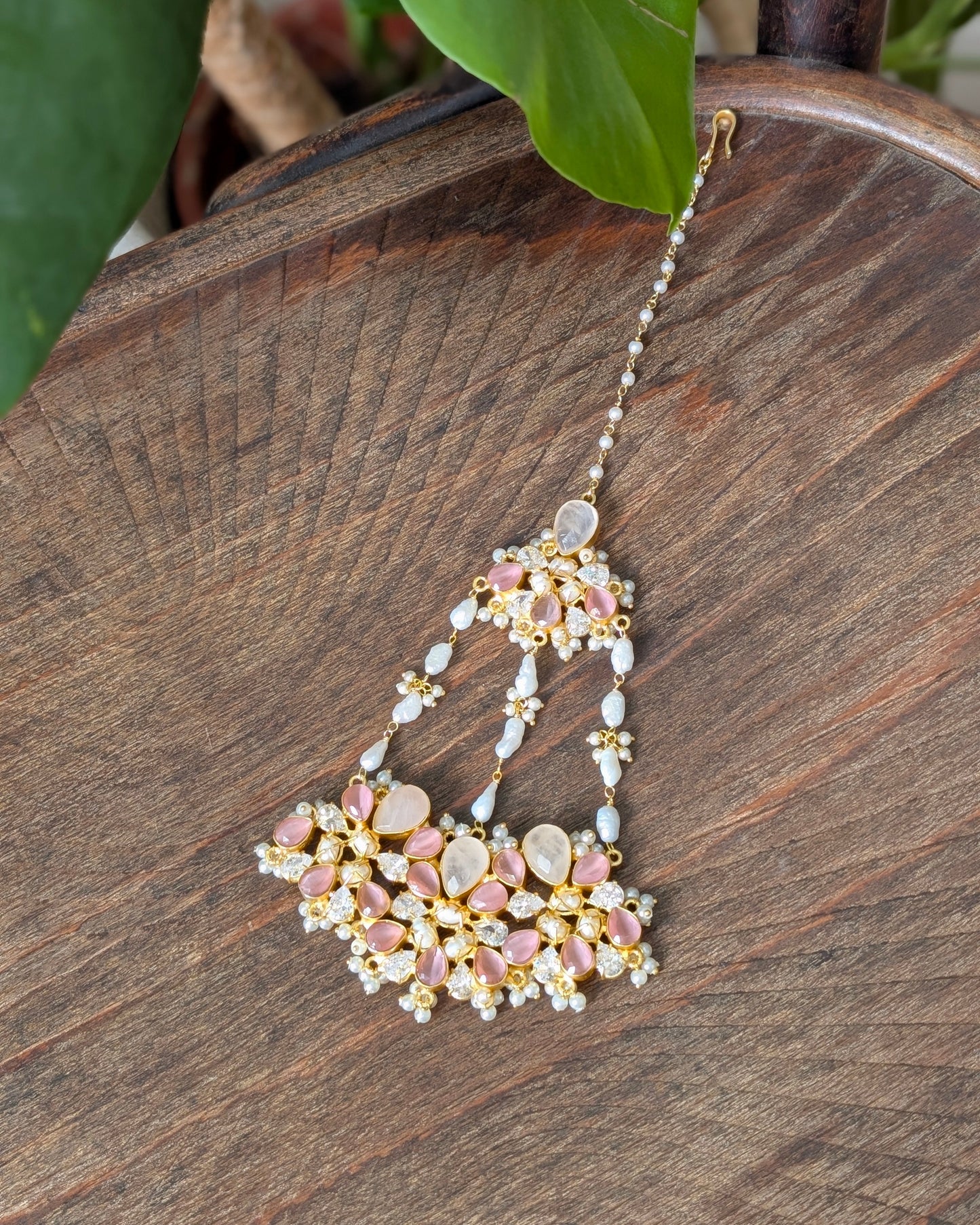 A detailed Indian style maang tikka with pink stones, white pearls, and gold-plated metal, resting on a dark wooden surface with green leaves in the background.