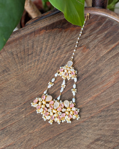 A detailed Indian style maang tikka with pink stones, white pearls, and gold-plated metal, resting on a dark wooden surface with green leaves in the background.