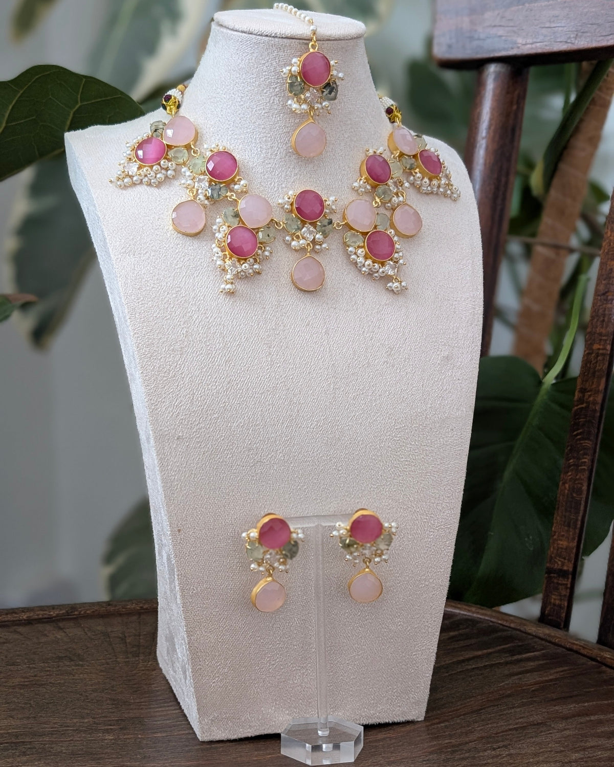 A gold-toned jewelry set with pink stones displayed on a cream jewelry bust, including a necklace, matching earrings, and a pair of drop earrings, set on a wooden table with green plants in the background.