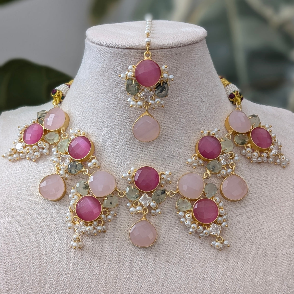 A gold-plated Indian ethnic jewelry set featuring a statement necklace, matching earrings, and a maang tikka, displayed on a cream-colored mannequin bust. The jewelry is decorated with round pink stones, small white pearls, and green enamel accents.