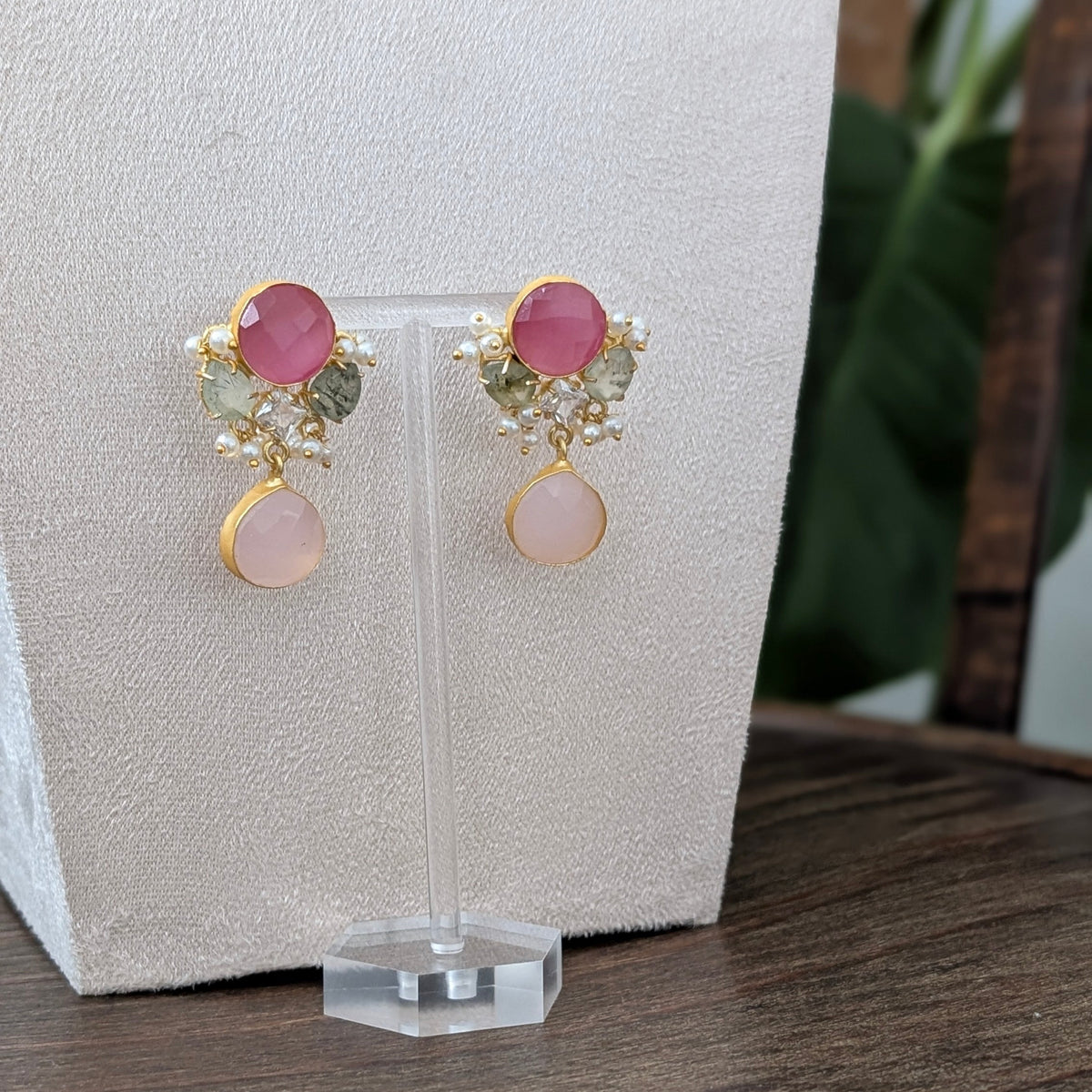 A pair of gold-toned dangle earrings with pink gemstones and small green leaf accents, displayed on a clear stand against a textured cream backdrop on a wooden surface.