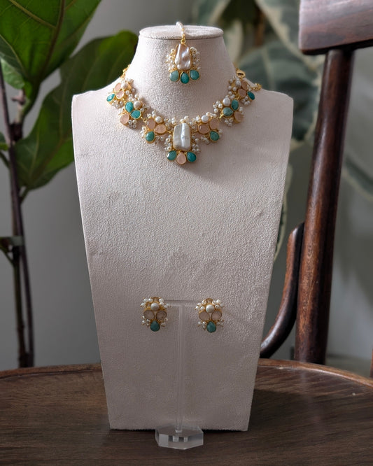 Gold-toned jewelry set displayed on a cream jewelry bust, including a necklace, matching earrings, and a small top piece, with turquoise green stones and pearl-like accents, set against a softly blurred indoor background with a plant and wooden chair.