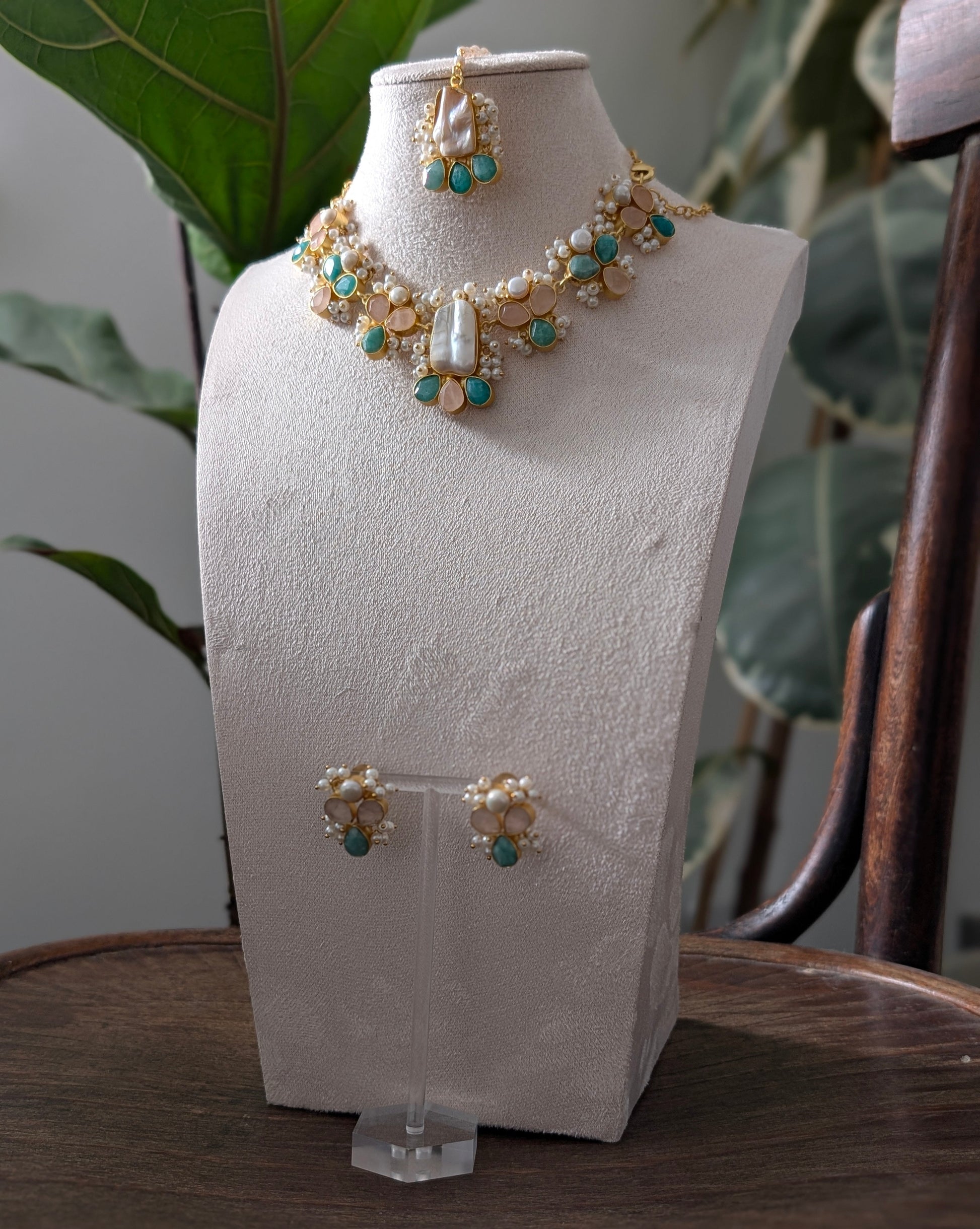 A gold and white jewelry set featuring a statement necklace and matching earrings, displayed on a textured white jewelry bust. The jewelry features white pearl-like beads, gold-toned metal, and turquoise blue stones, set against a background of green houseplants.