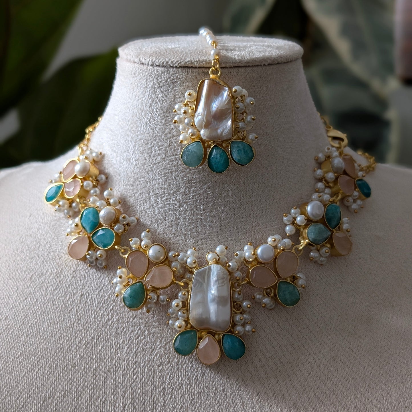 Gold-plated statement necklace and matching maang tikka set with turquoise blue stones, white pearls, and peach-colored beads, displayed on a beige velvet jewelry bust.