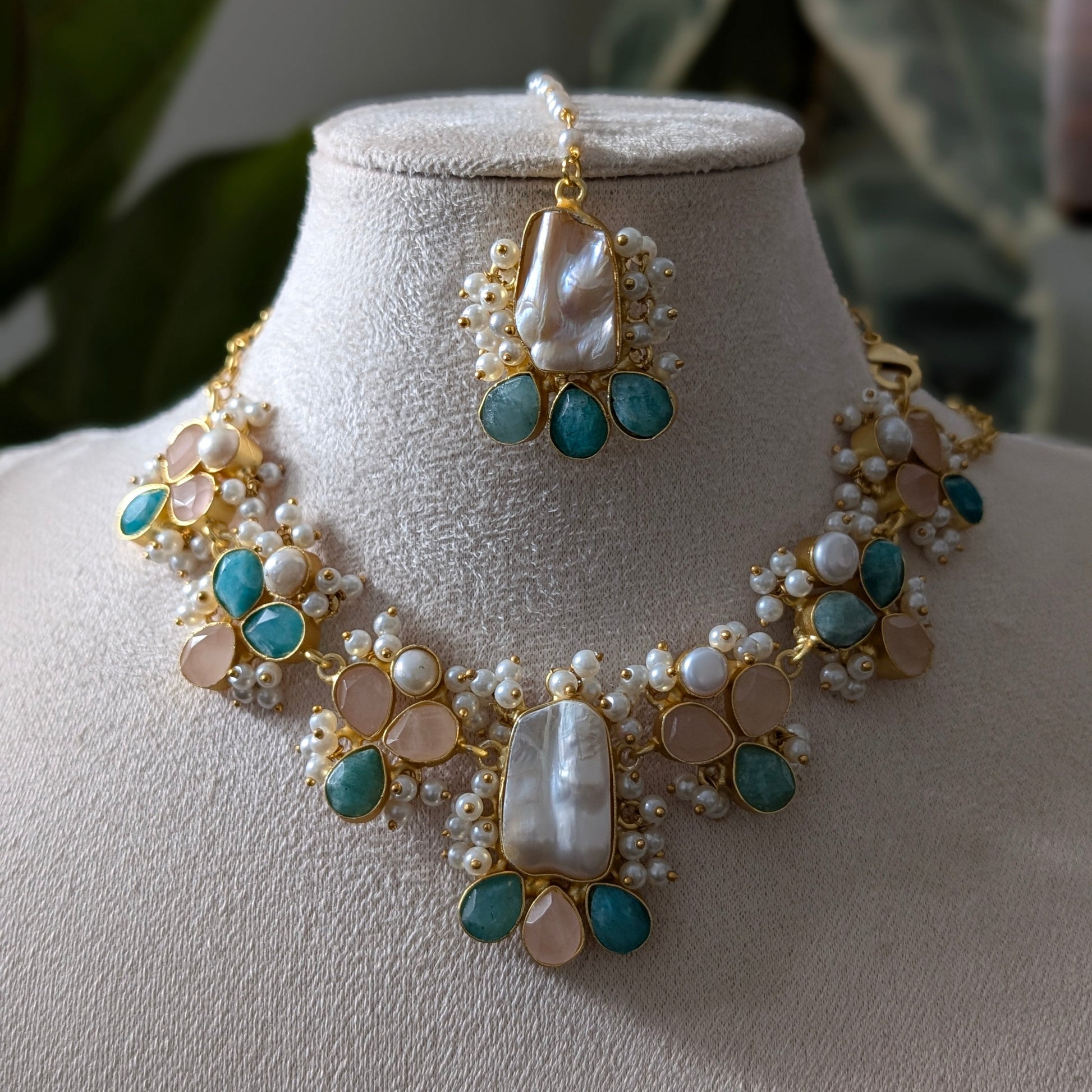 Gold-plated statement necklace and matching maang tikka set with turquoise blue stones, white pearls, and peach-colored beads, displayed on a beige velvet jewelry bust.