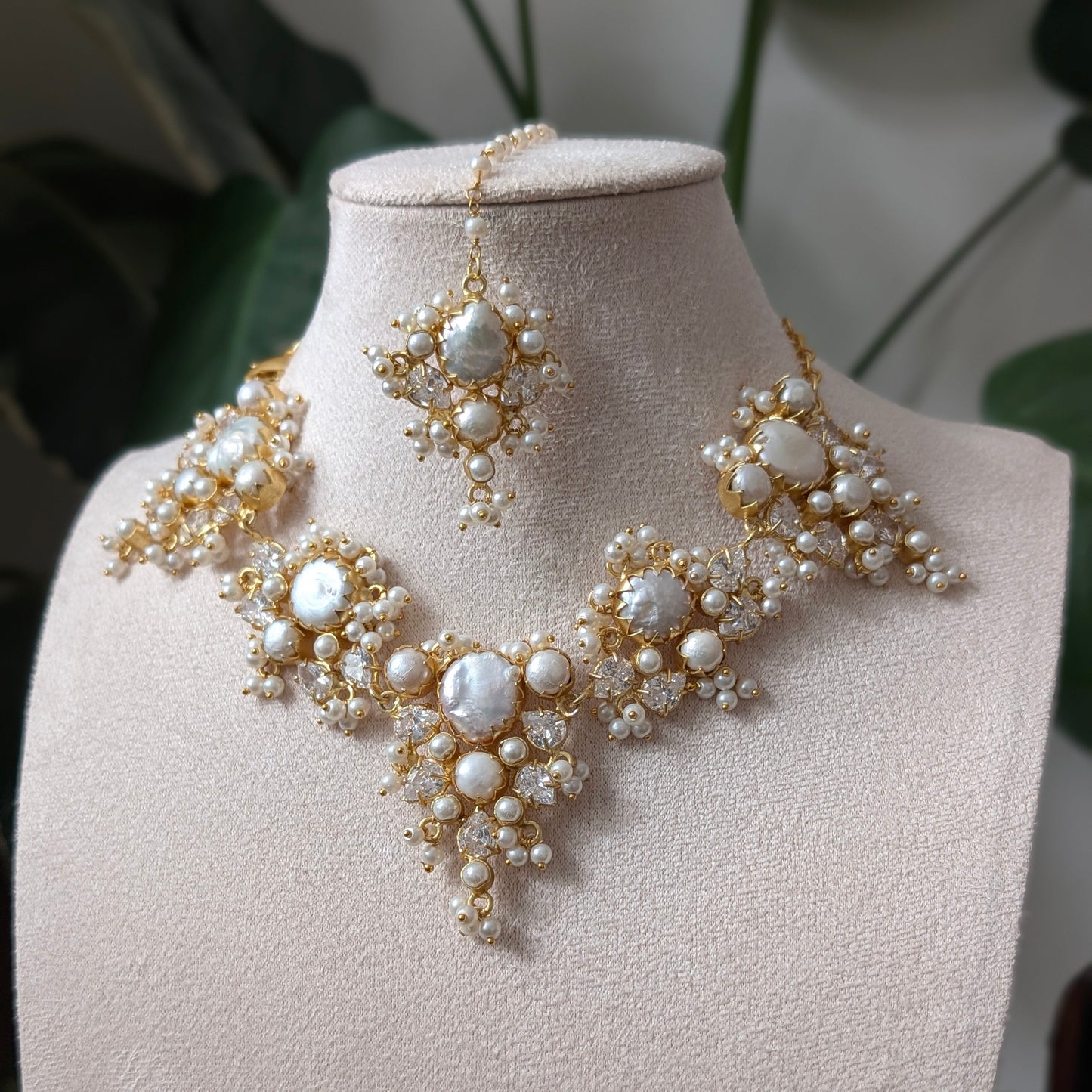 Gold and pearl jewelry set displayed on a beige jewelry bust, featuring a statement necklace, matching earrings, and a maang tikka, with a soft-focus background of green leaves.