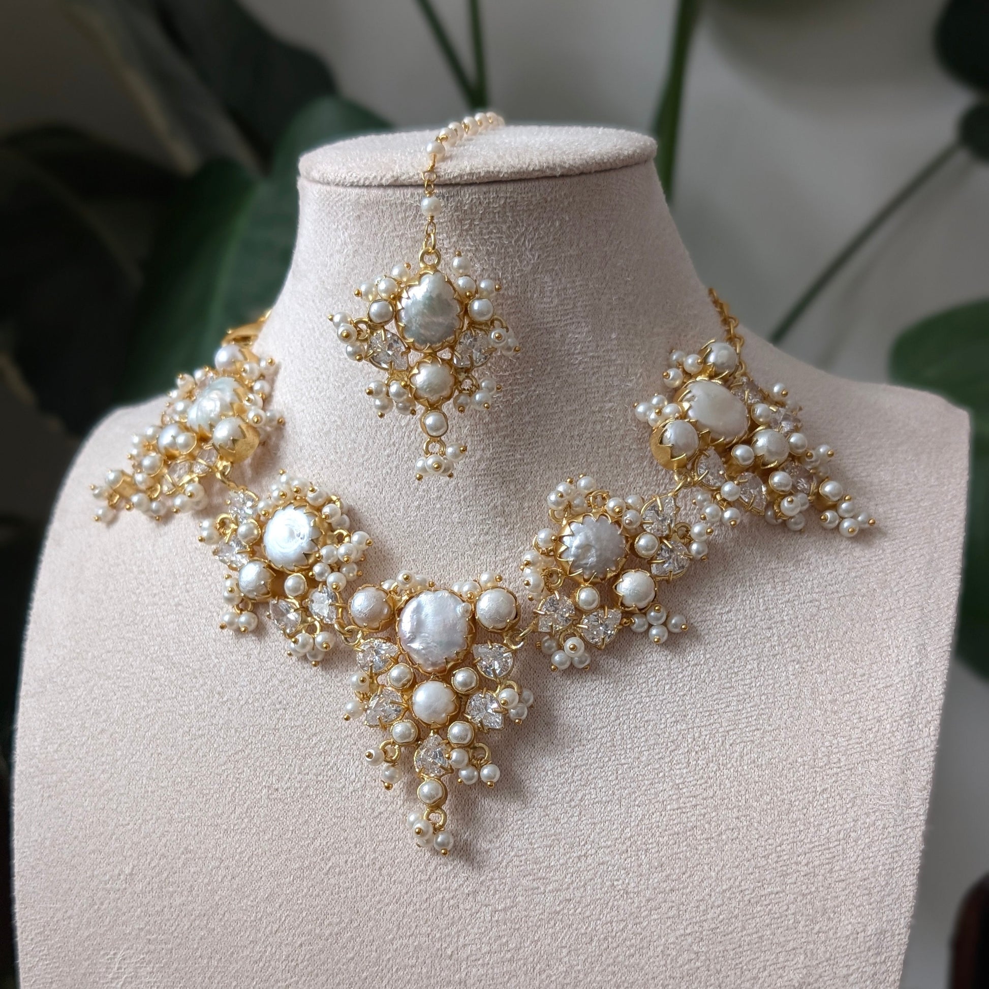 Gold and pearl jewelry set displayed on a beige jewelry bust, featuring a statement necklace, matching earrings, and a maang tikka, with a soft-focus background of green leaves.