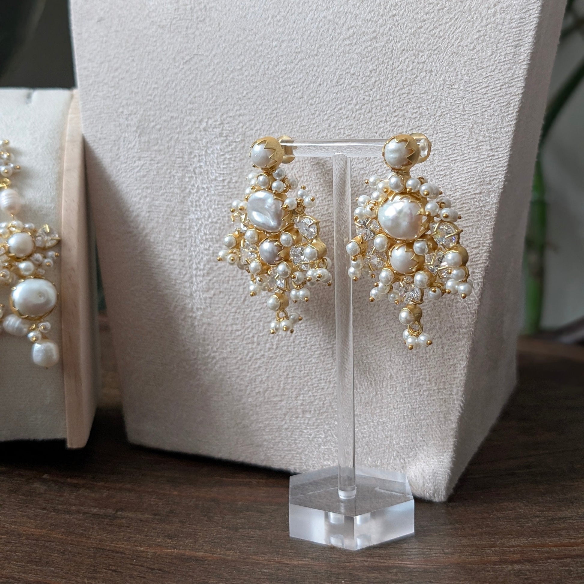 Gold and white pearl statement drop earrings displayed on a clear acrylic stand in front of a cream jewelry bust, with a matching display box partially visible on the left.