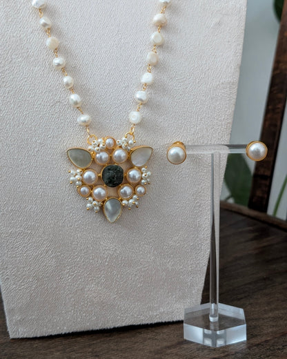 A gold and pearl necklace with a central dark green stone pendant, displayed on a cream fabric jewelry bust alongside matching pearl stud earrings on a clear stand.