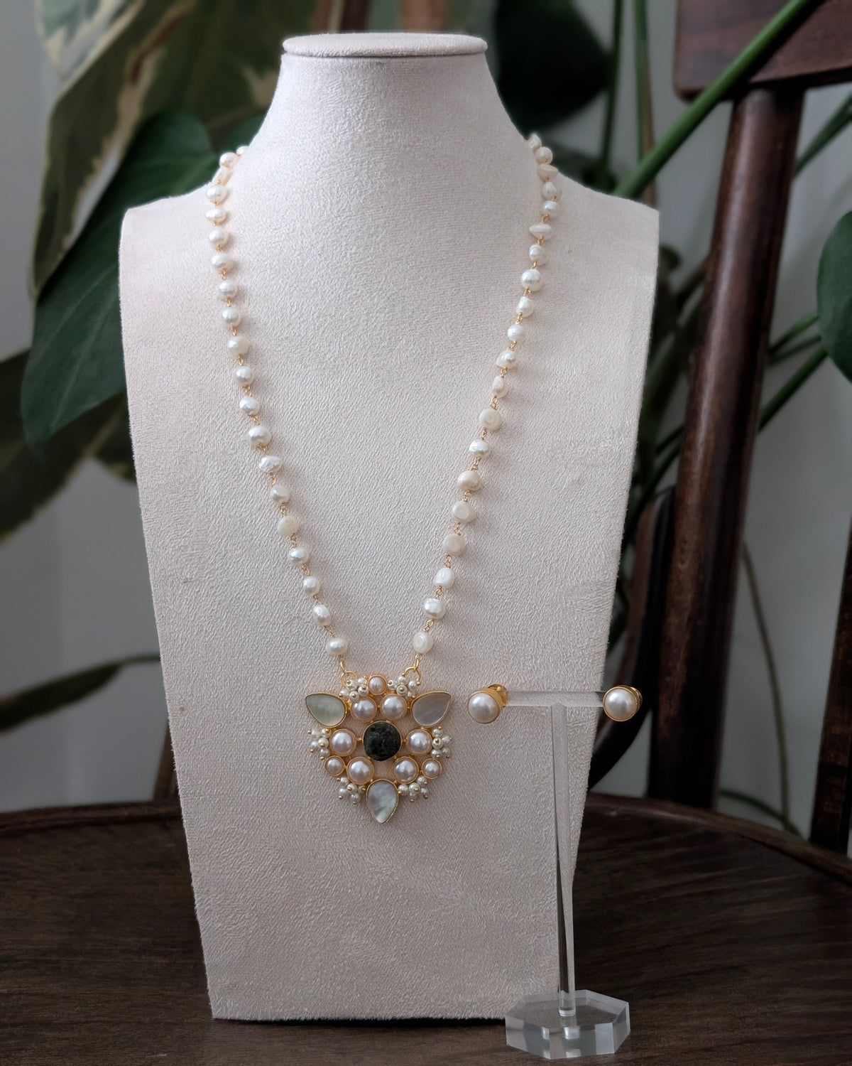 A pearl necklace with a gold-toned pendant and matching pearl stud earrings displayed on a cream fabric mannequin bust, set on a dark wooden table with green foliage in the background.