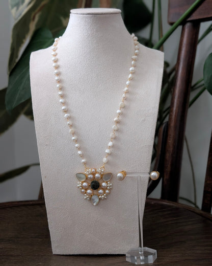 A pearl necklace with a gold-toned pendant and matching pearl stud earrings displayed on a cream fabric mannequin bust, set on a dark wooden table with green foliage in the background.