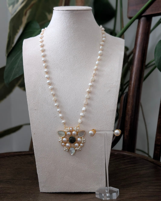 A pearl necklace with a gold-toned pendant and matching pearl stud earrings displayed on a cream fabric mannequin bust, set on a dark wooden table with green foliage in the background.
