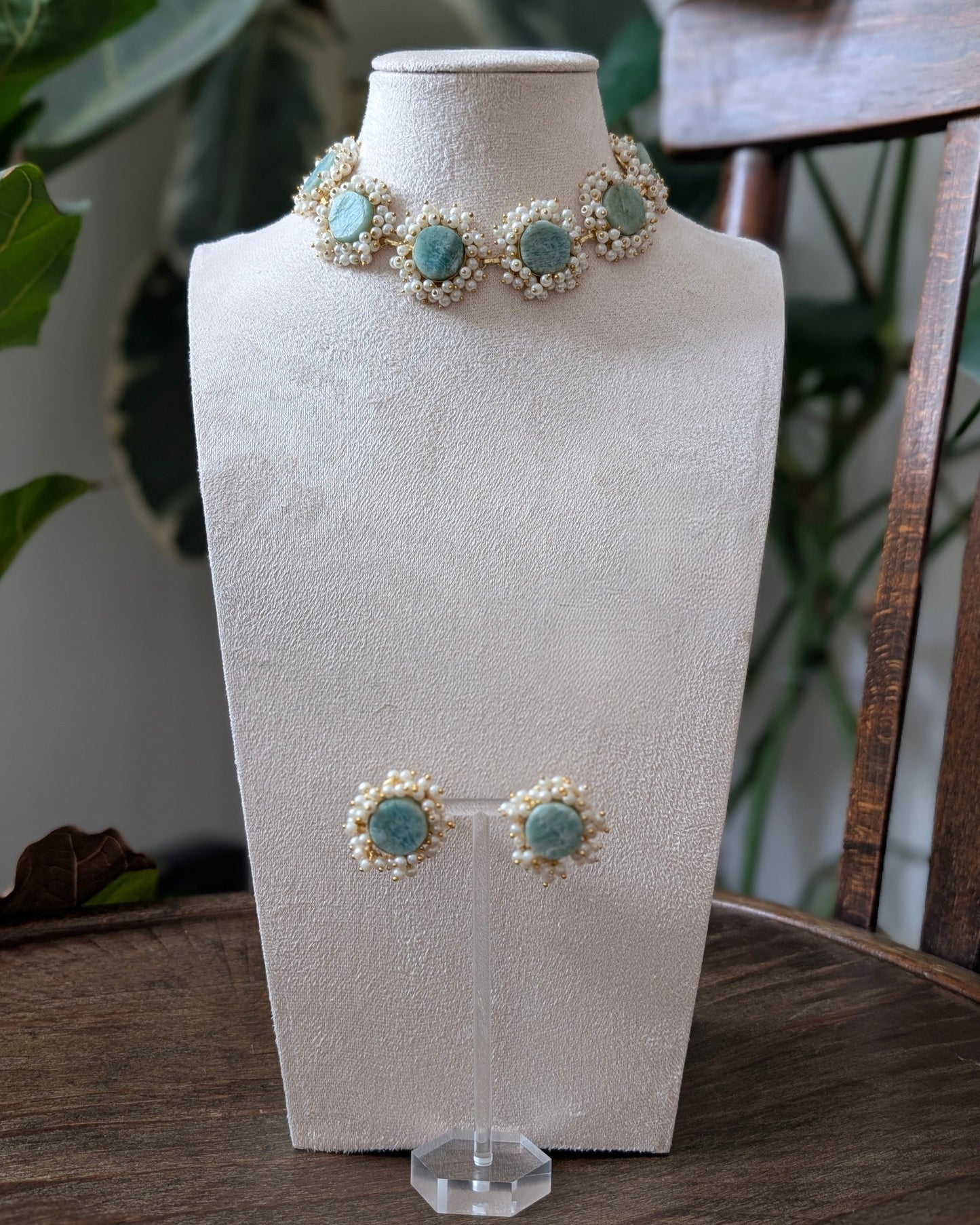 A jewelry set displayed on a cream fabric mannequin bust, featuring a pearl and turquoise necklace with matching stud earrings, set on a wooden table with a chair and plants in the background.